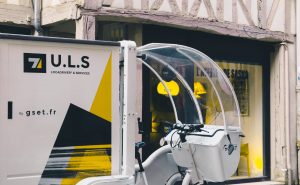 Vélo-cargo électrique blanc GSET, stationné dans une rue pavée de Rouen. Le véhicule est équipé d'une remorque fermée et arbore les logos U.L.S et GSET. Livraison urbaine écologique et flexible.