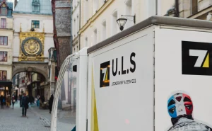 Vélo-cargo électrique U.L.S stationné devant la grande horloge de Rouen. Le véhicule est garé sur une place animée, prêt pour une nouvelle livraison. Solution de livraison urbaine écologique et flexible au cœur de la ville historique.