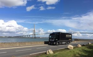Camion de livraison E.U.RO roulant sur une route côtière, avec le pont de Normandie en arrière-plan. Transport routier de marchandises en France.